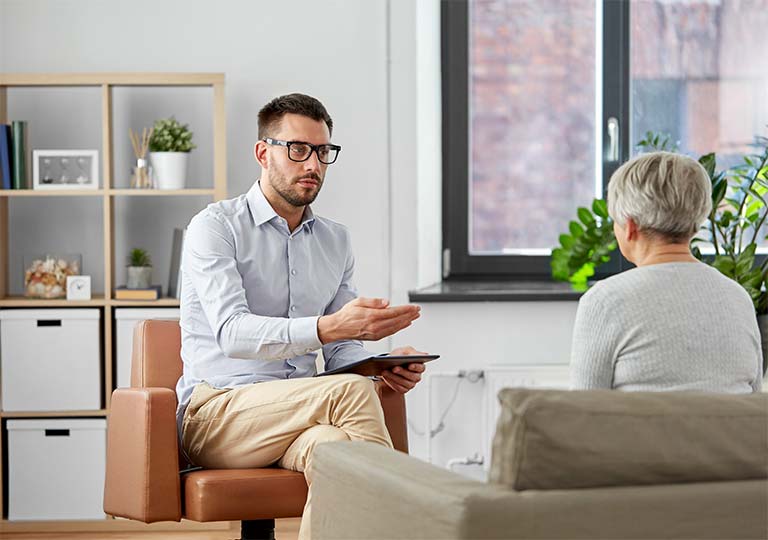 A man with glasses sits in a chair holding a tablet and talking to an older woman seated opposite him in a room with modern decor.