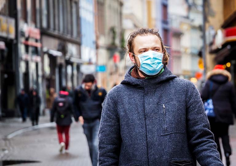 A man wearing a face mask and a blue coat walks down a busy urban street. Other people in the background are also wearing face masks. Buildings line both sides of the street.