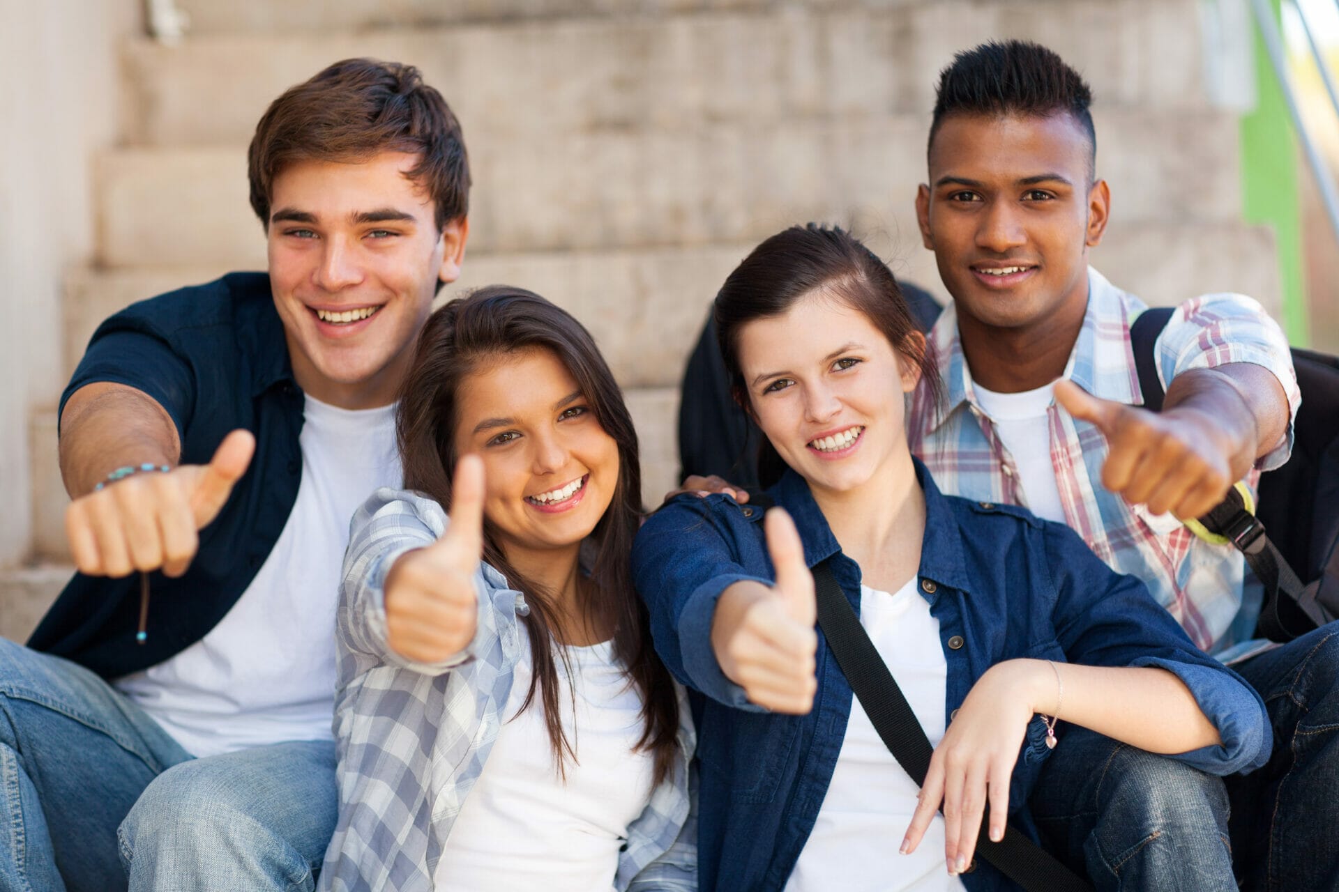 Cuatro personas sonrientes, sentadas en unas escaleras al aire libre, hacen un gesto con el pulgar hacia arriba ante la cámara. Están vestidas con ropa informal y parecen felices y relajadas, encarnando el espíritu de cuidado de los adolescentes.