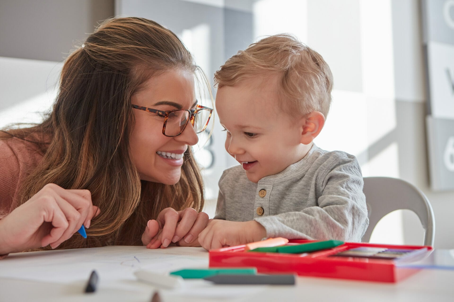Un adulto y un niño dibujan en una mesa, fomentando la creatividad y el vínculo. El adulto lleva gafas y ambos sonríen. En la mesa se ven herramientas de dibujo y papel, lo que resalta sutilmente la importancia de las evaluaciones del desarrollo a través de su alegre interacción.
