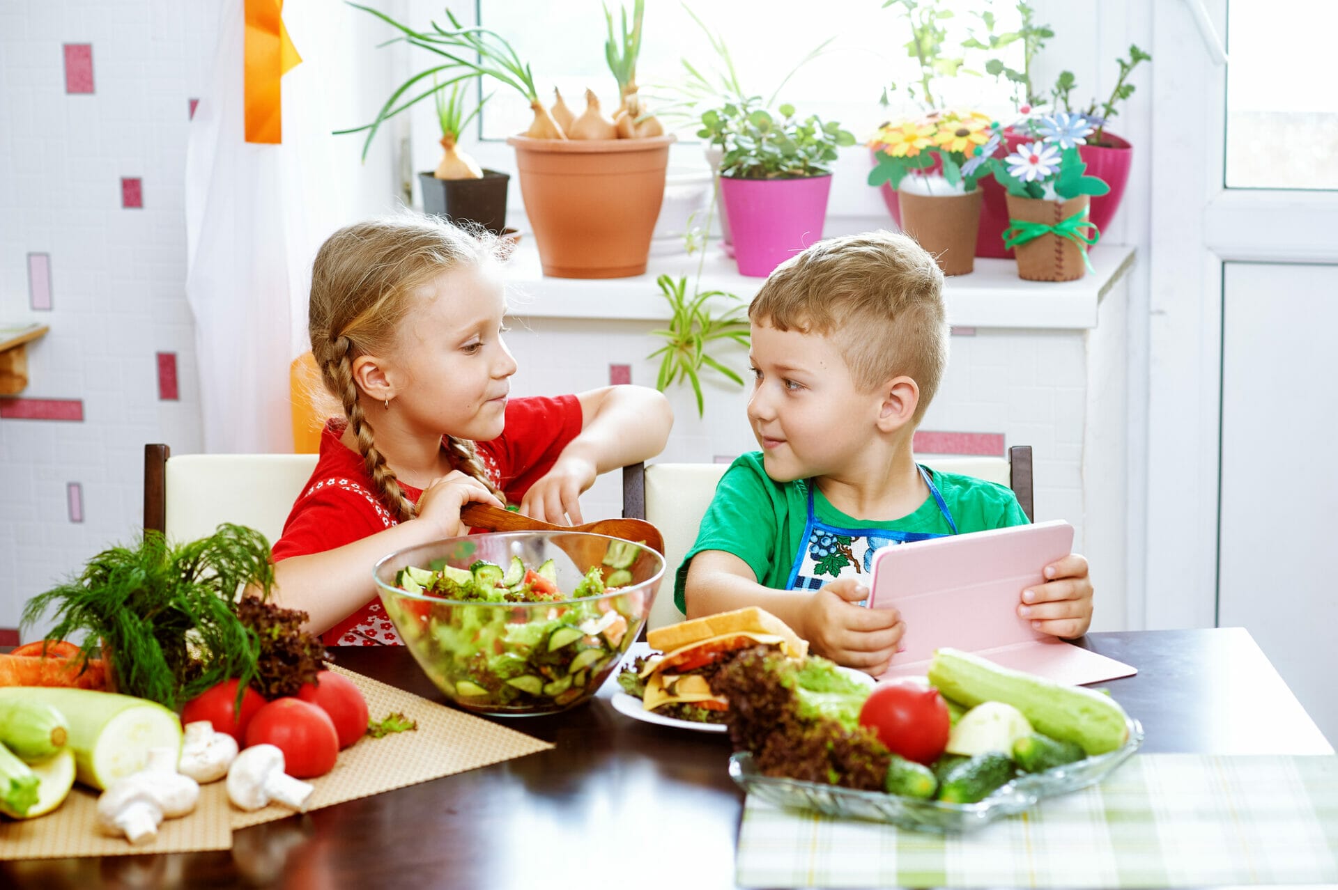 Dos niños están sentados a una mesa con diversas verduras y ensalada. Uno prepara una ensalada nutritiva mientras el otro sostiene una tableta. En el alféizar de la ventana, al fondo, hay plantas en macetas.