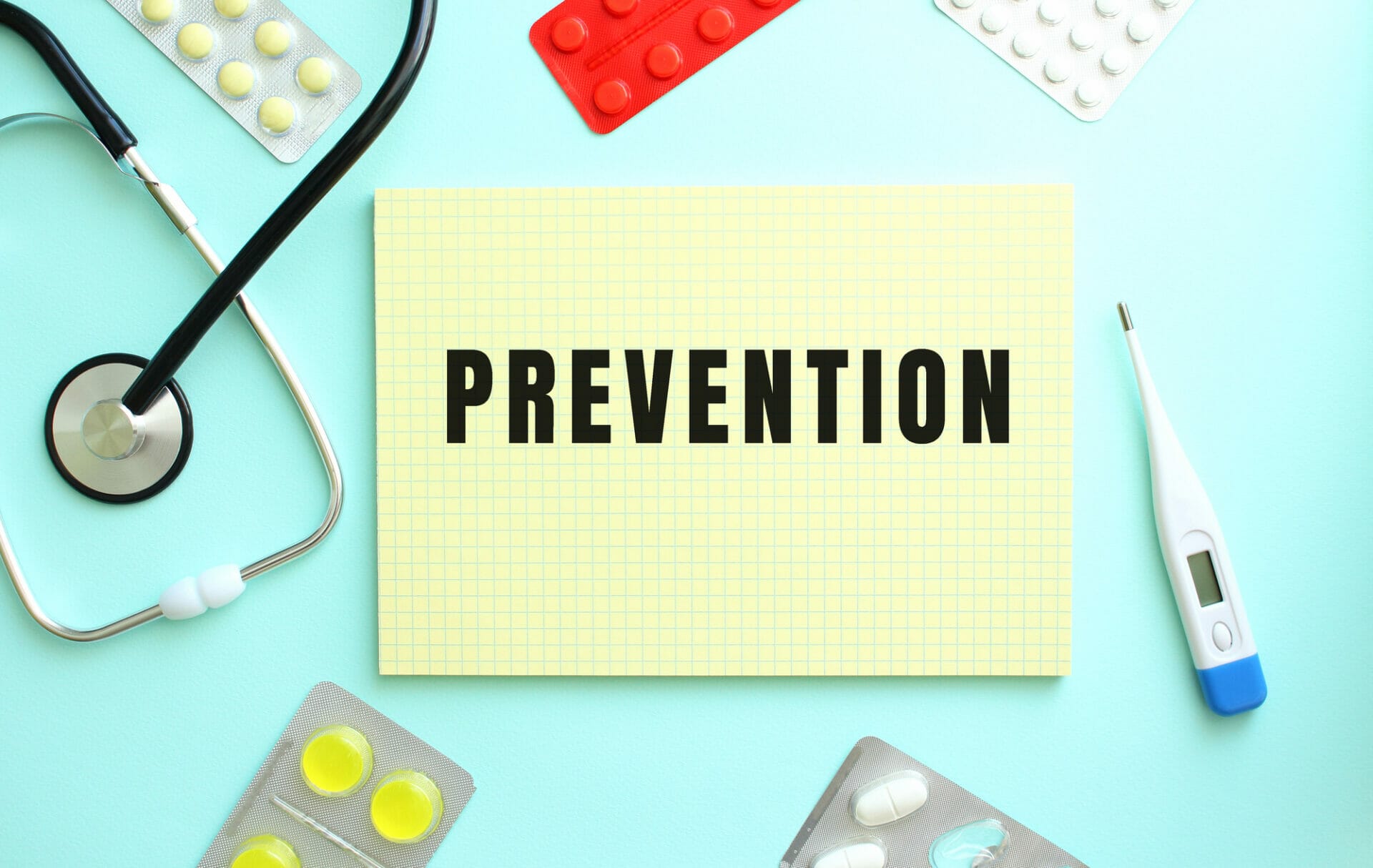 Un bloc de notas amarillo con la palabra "PREVENCIÓN", rodeado de artículos médicos, entre ellos un estetoscopio, un termómetro y varios blísters de pastillas, enfatiza la importancia de la salud preventiva sobre un fondo azul claro.
