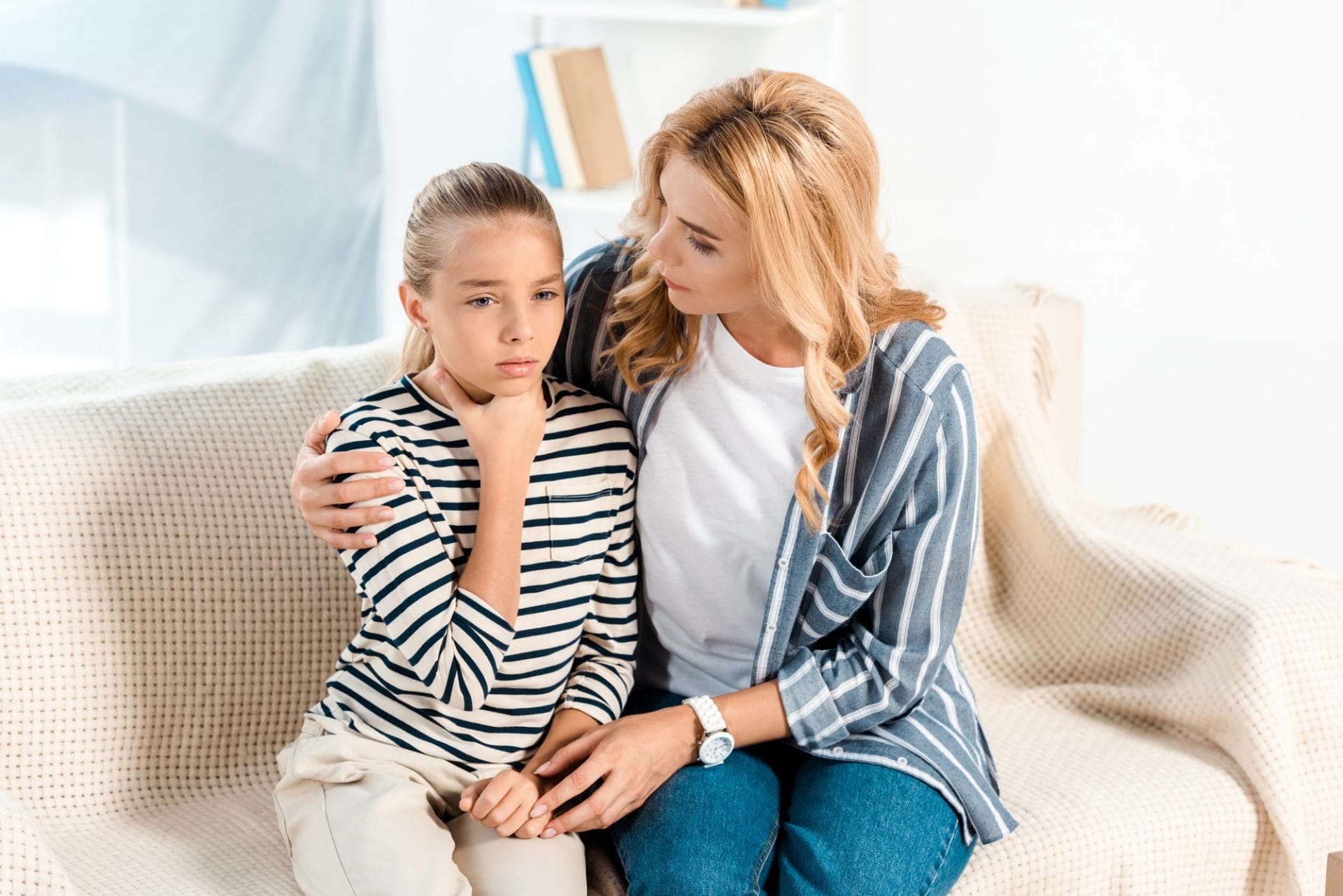 Una mujer está sentada en un sofá con su hija, increíblemente preocupada y agarrándose la garganta de dolor, buscando ayuda desesperadamente.