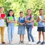 Cinco niños están al aire libre, con libros en la mano y sonriendo a la cámara. Vestidos de manera informal y con mochilas, disfrutan de un saludable regreso a la escuela en medio del follaje otoñal, con árboles y vegetación de fondo.