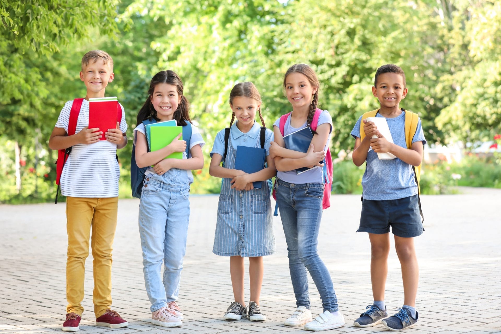 Cinco niños están al aire libre, con libros en la mano y sonriendo a la cámara. Vestidos de manera informal y con mochilas, disfrutan de un saludable regreso a la escuela en medio del follaje otoñal, con árboles y vegetación de fondo.