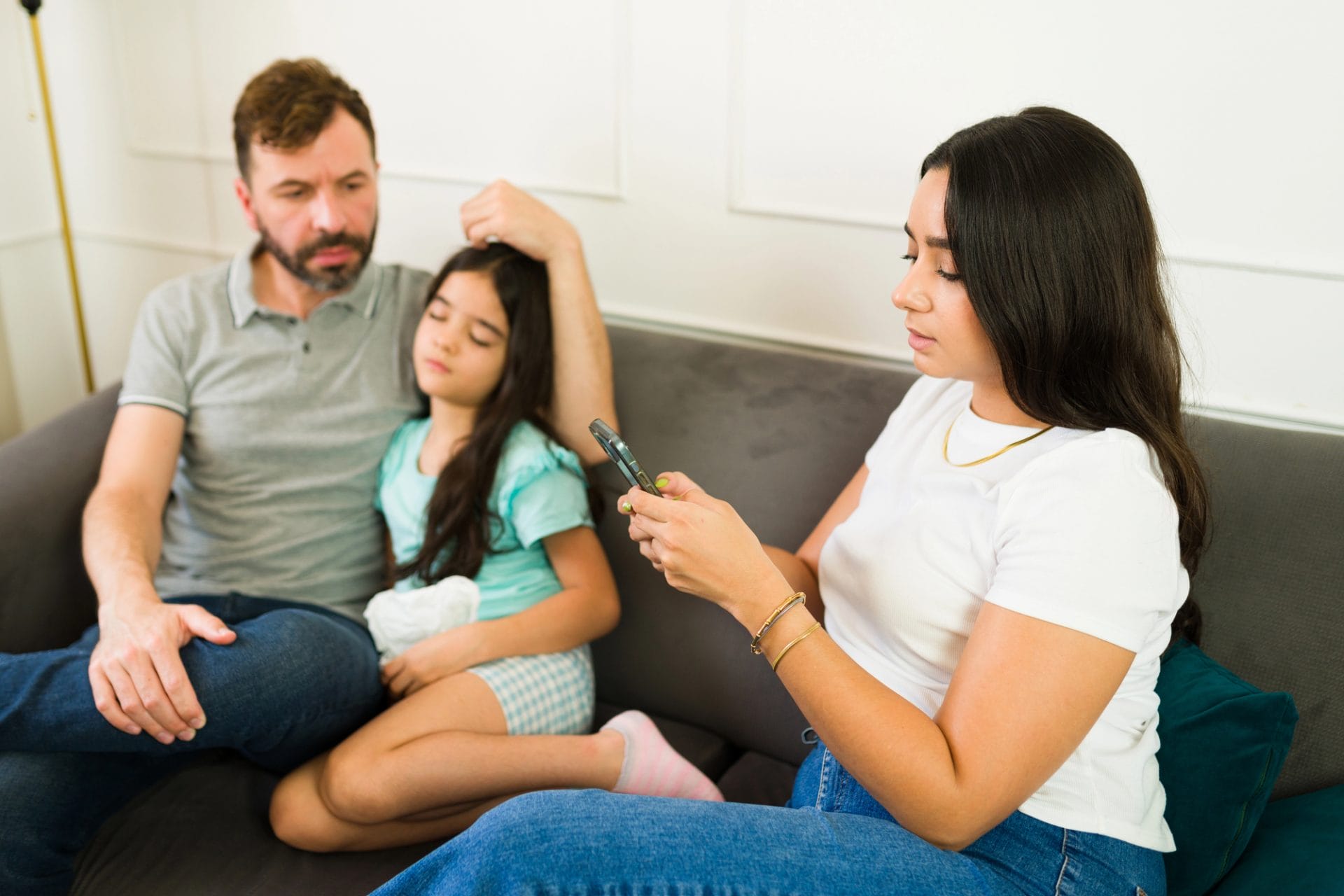 Un hombre y una niña están sentados juntos en un sofá, mientras una mujer a su lado se concentra en su teléfono inteligente.