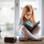 Una niña con gafas, impulsada por la curiosidad infantil, está sentada con las piernas cruzadas sobre una mesa de madera, absorta en su libro. Lápices de colores y más libros la rodean, alimentando su imaginación.