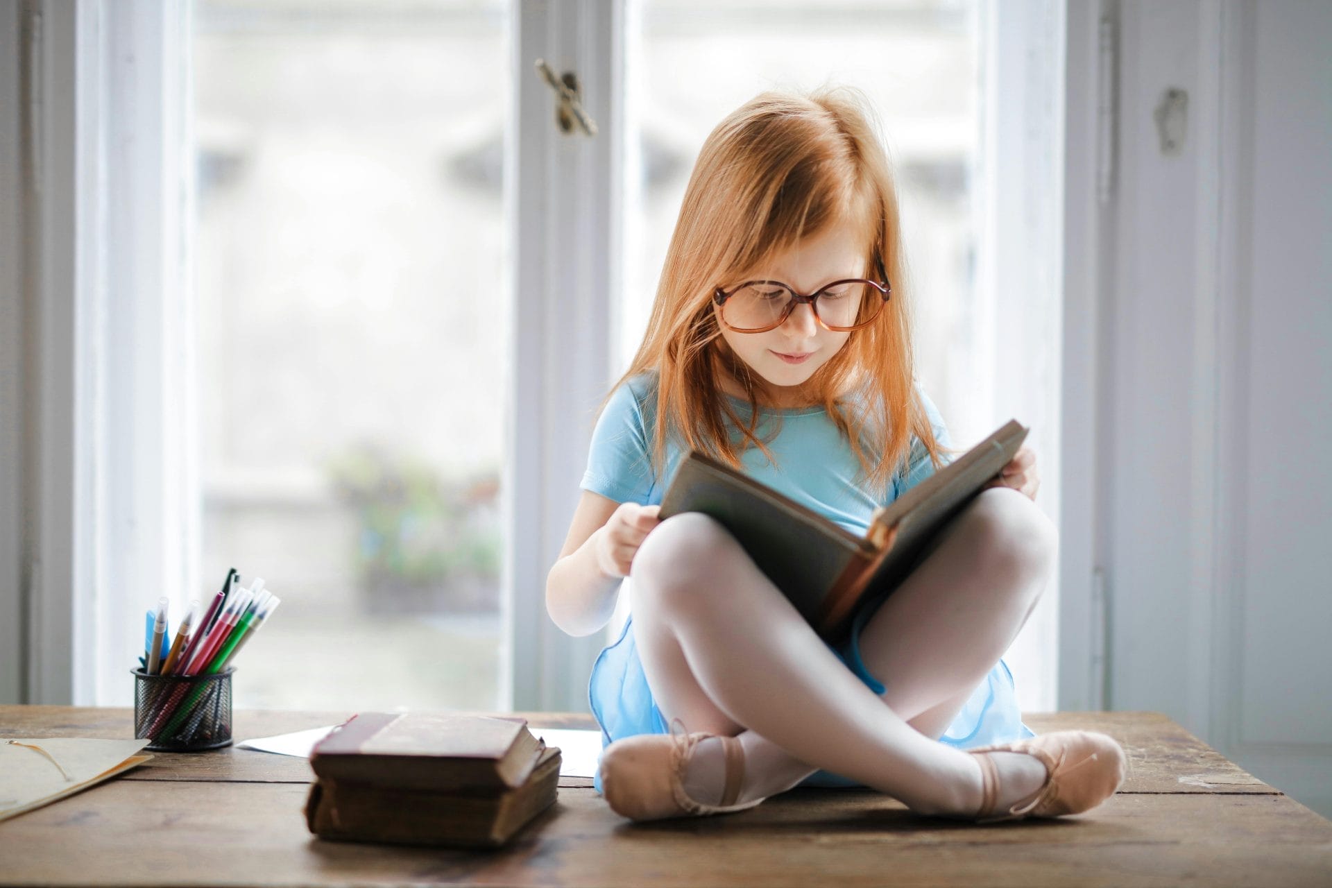 Una niña con gafas, impulsada por la curiosidad infantil, está sentada con las piernas cruzadas sobre una mesa de madera, absorta en su libro. Lápices de colores y más libros la rodean, alimentando su imaginación.