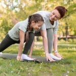Una mujer y una niña practican yoga sobre una colchoneta en el parque, ambas en posición de estocada y sonriendo, adoptando el autocuidado familiar para un hogar más saludable.