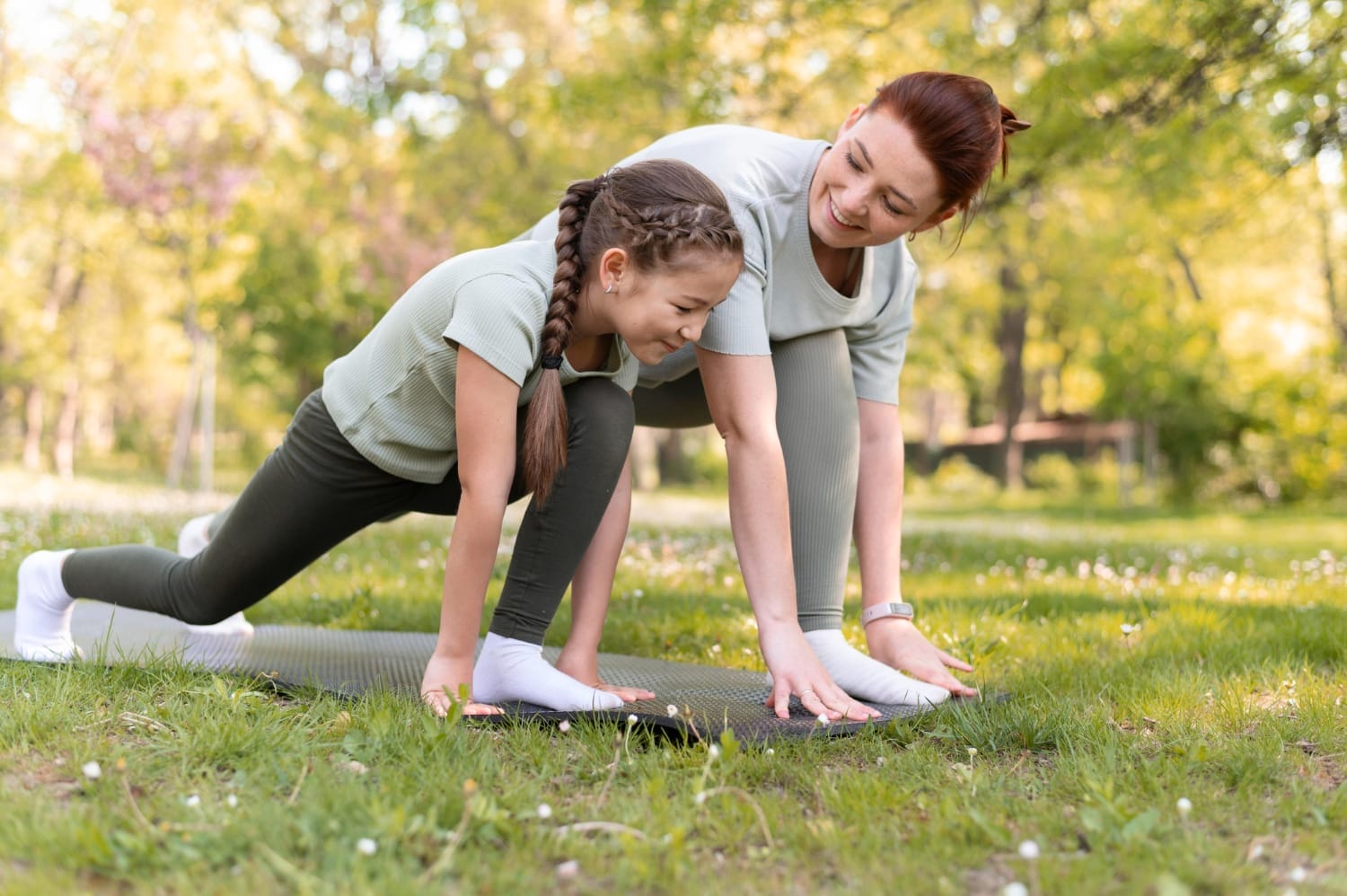Una mujer y una niña practican yoga sobre una colchoneta en el parque, ambas en posición de estocada y sonriendo, adoptando el autocuidado familiar para un hogar más saludable.