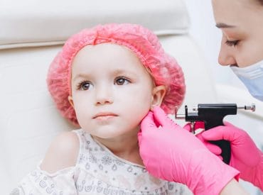En el suroeste de Houston, un niño pequeño con una redecilla rosa para el pelo recibe una perforación en la oreja de un profesional enmascarado con guantes rosas, lo que garantiza la seguridad y la precisión, un testimonio de la calidad de los servicios de perforación de orejas pediátricas.