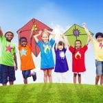 Seis niños que visten camisetas coloridas con símbolos están parados en el césped, sosteniendo cometas y sonriendo bajo un cielo azul, mostrando cómo las familias pueden ayudar a sus hijos a tomar decisiones saludables a través de divertidas actividades al aire libre.