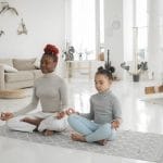 Two people, an adult and a child, sit cross-legged on a yoga mat in a bright living room, meditating with their eyes closed—a peaceful moment of teaching kids self-care through mindful parenting.