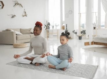 Two people, an adult and a child, sit cross-legged on a yoga mat in a bright living room, meditating with their eyes closed—a peaceful moment of teaching kids self-care through mindful parenting.
