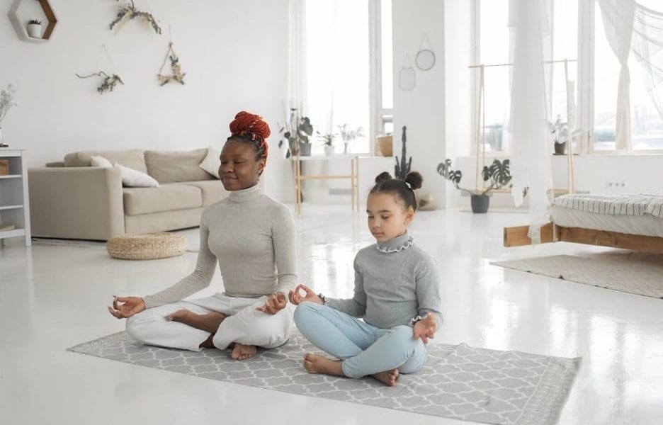 Two people, an adult and a child, sit cross-legged on a yoga mat in a bright living room, meditating with their eyes closed—a peaceful moment of teaching kids self-care through mindful parenting.