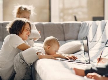 A woman sits on a couch with two small children; one baby leans over to touch a laptop keyboard, illustrating hands-on parenting while trying to raise busy kids.