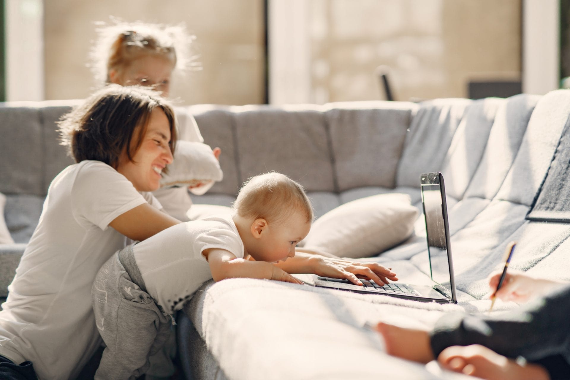 A woman sits on a couch with two small children; one baby leans over to touch a laptop keyboard, illustrating hands-on parenting while trying to raise busy kids.