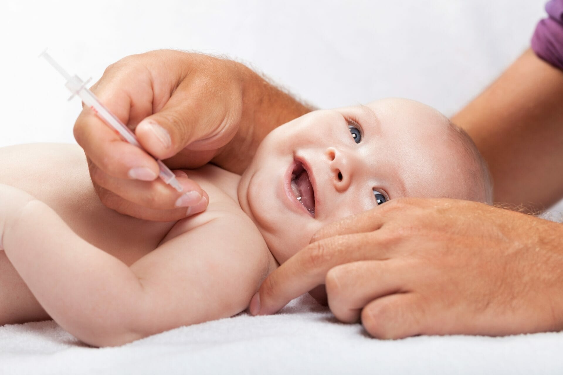 A smiling baby lies down while an adult hand holds a syringe, seemingly administering vaccinations in the baby's arm.