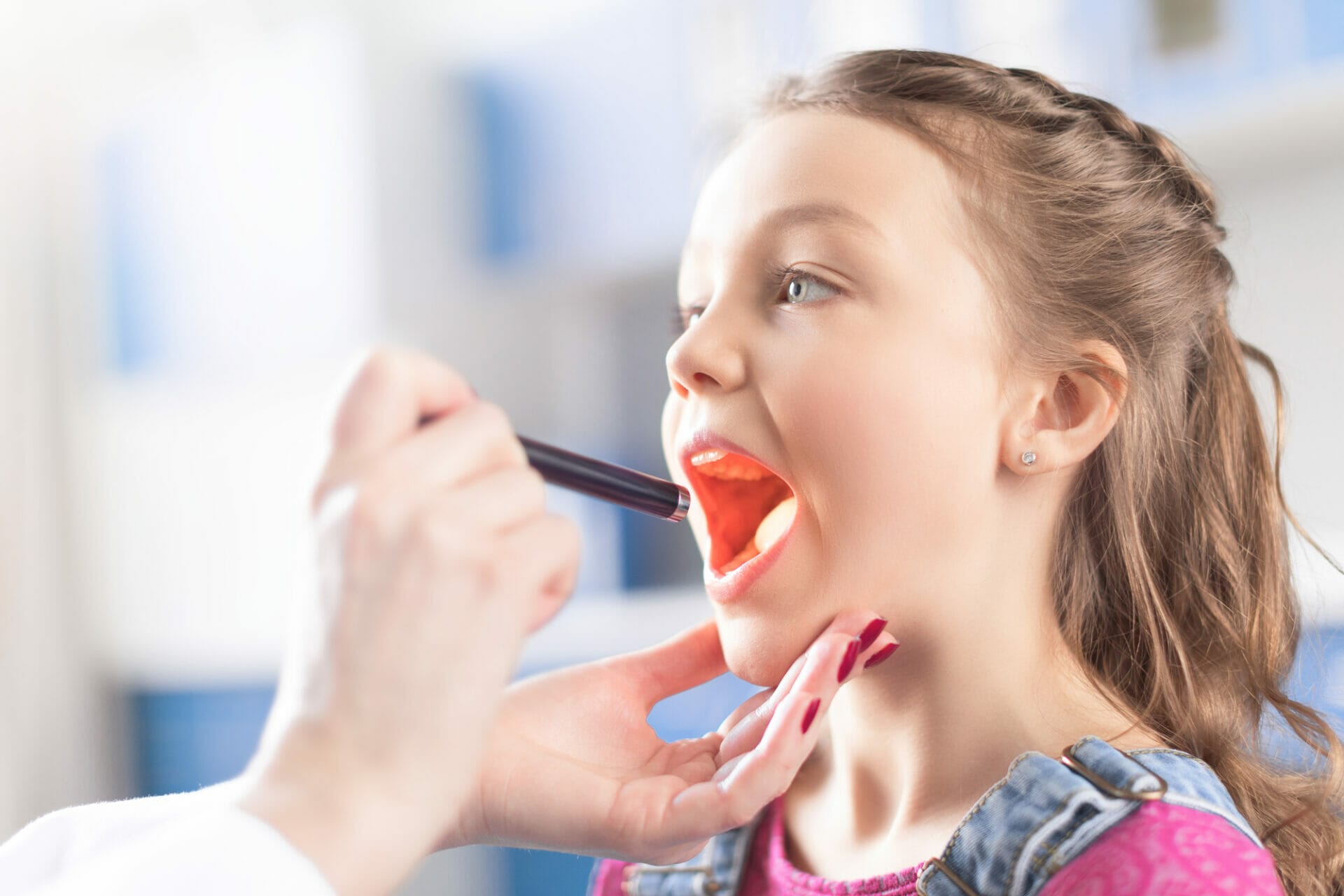 A young girl in a pink shirt opens her mouth wide as a healthcare professional examines her throat with a penlight, ensuring she's healthy and ready for vaccinations.