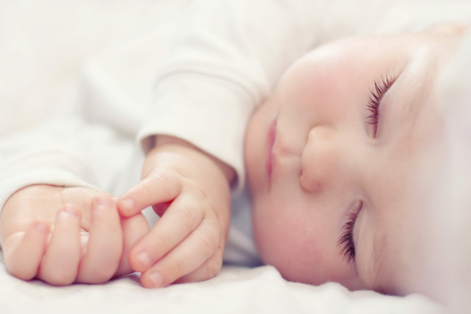 Close-up of a sleeping baby with closed eyes, long eyelashes, and hands resting near the face, wearing a white outfit—a peaceful image reminding us that proper adolescent care starts from nurturing these tender moments.