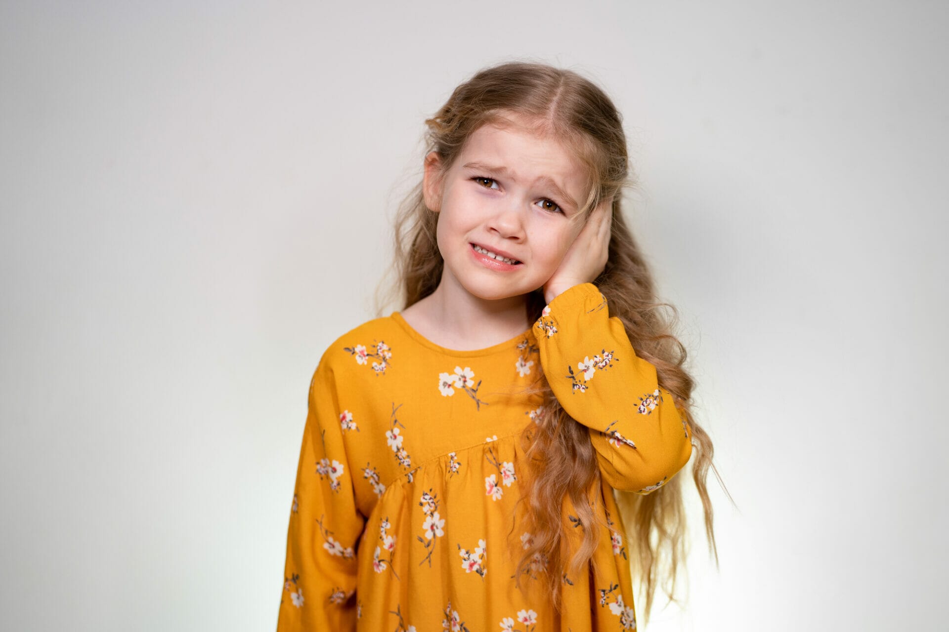 A young girl with long wavy hair, wearing a yellow floral dress, holds her hand to her temple with a concerned expression, possibly indicating an earache.