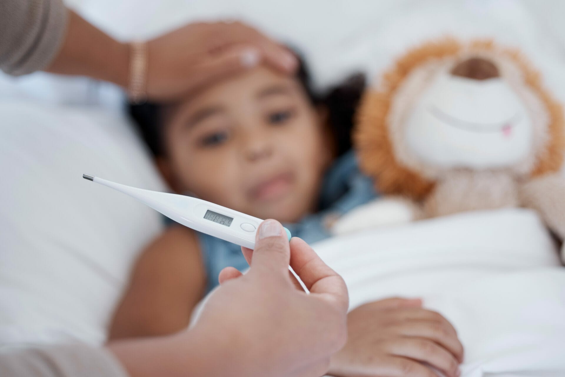 A person holds a thermometer while another touches a child's forehead; the child, showing signs of fever, lies in bed with a stuffed lion toy.