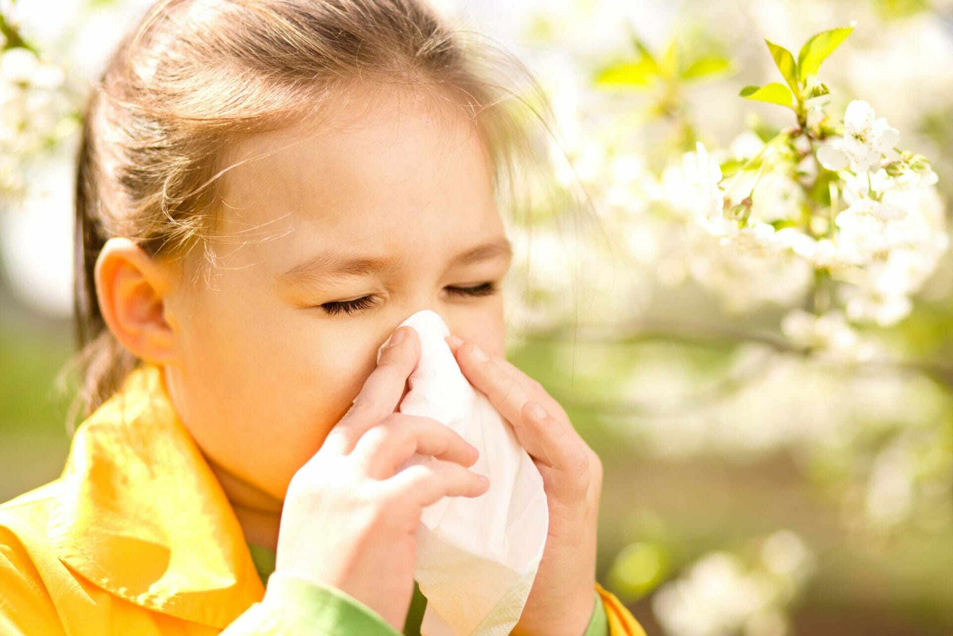A young girl in a yellow jacket sneezes into a tissue while standing outdoors among blooming flowers, clearly battling allergies.