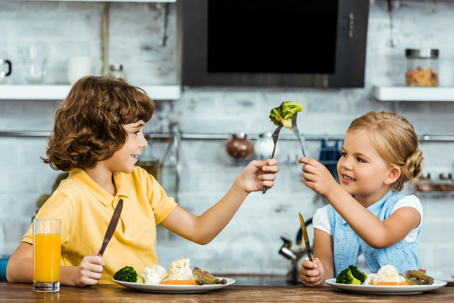 Two children sit at a table with plates of vegetables, playfully holding a fork with broccoli towards each other. A glass of orange juice reinforces the scene's focus on nutrition, sitting in front of one child. The kitchen is in the background.