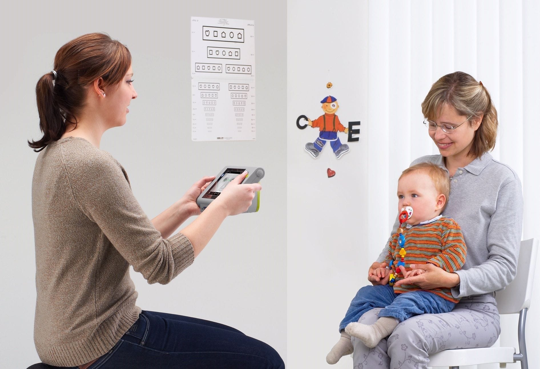 A healthcare worker conducts an early detection eye test on a young child, who is held by an adult woman, utilizing digital vision screening technology in a clinical setting.