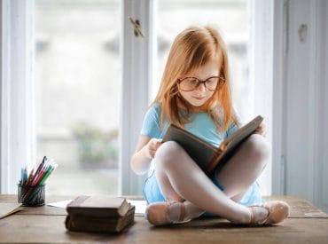 A young girl with glasses, driven by a child's curiosity, sits cross-legged on a wooden table, engrossed in her book. Colored pencils and more books surround her, nurturing her imagination.