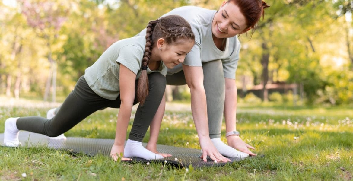 Una mujer y una niña practican yoga sobre una colchoneta en el parque, ambas en posición de estocada y sonriendo, adoptando el autocuidado familiar para un hogar más saludable.