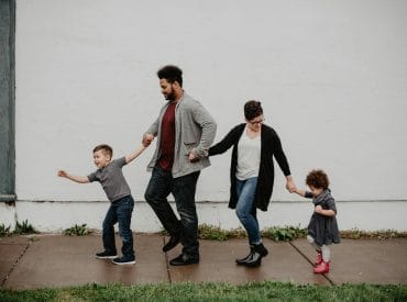 A family of four, embodying modern parenting, walks hand in hand along a sidewalk before a white wall, with the children on the outside and parents in the middle.