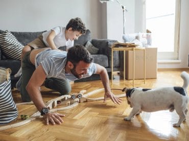 An adult crawls on the floor with a child on his back, enjoying indoor adventures with a toy train set, while a small dog stands nearby in a living room filled with boxes and a sofa. An adult crawls on the floor with a child on his back, enjoying indoor adventures with a toy train set, while a small dog stands nearby in a living room filled with boxes and a sofa.