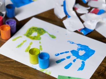 A sheet of paper with blue and green handprints next to open paint pots, surrounded by torn paper scraps on a wooden surface—perfect for fun memories and rainy day kids projects. A sheet of paper with blue and green handprints next to open paint pots, surrounded by torn paper scraps on a wooden surface—perfect for fun memories and rainy day kids projects.